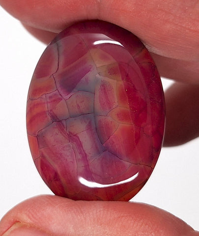Oval-shaped gemstone with red and purple patterns held between fingers